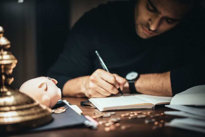 A man writing in an account book by lamplight, coins and a piggy bank on the desk